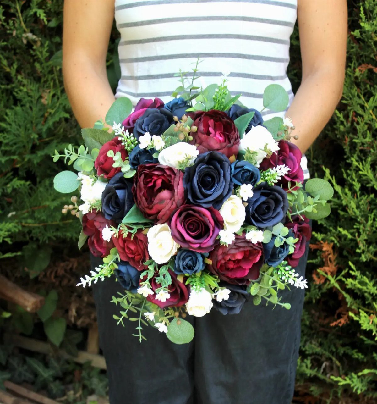 A bouquet of burgundy, navy blue, and white roses with green eucalyptus leaves held by a person.
