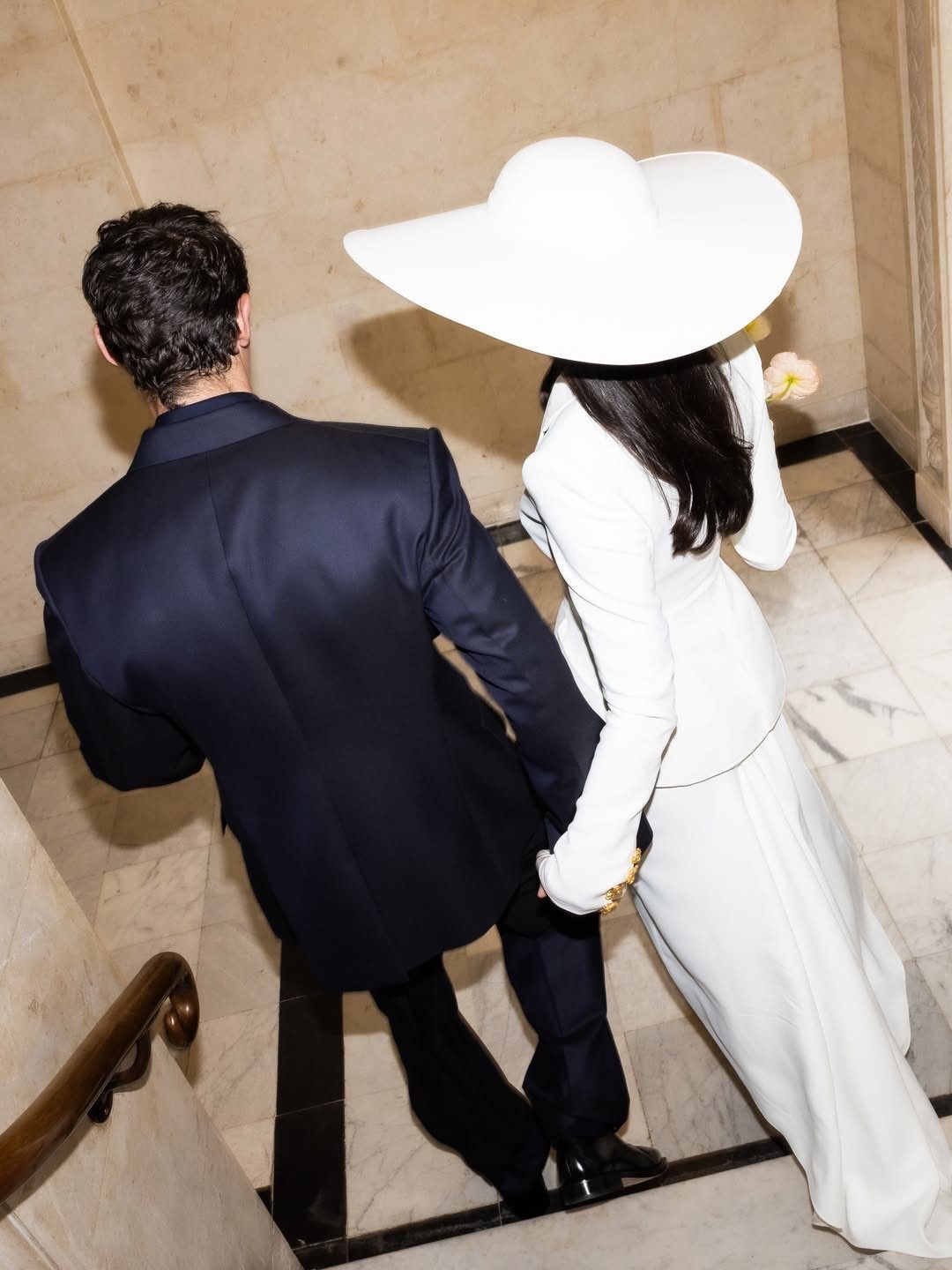 A man in a navy suit and a woman in a white suit and large hat walking up stairs.