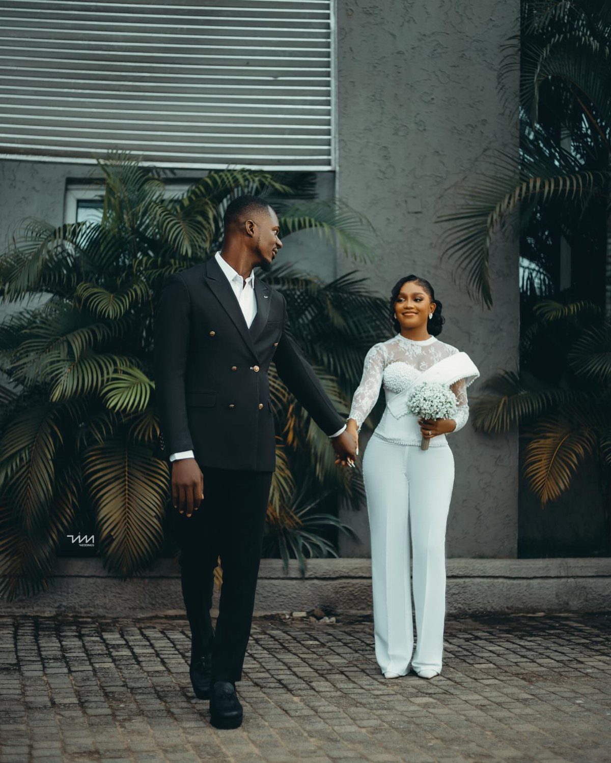 A bride in a white lace jumpsuit and a groom in a black suit holding hands.