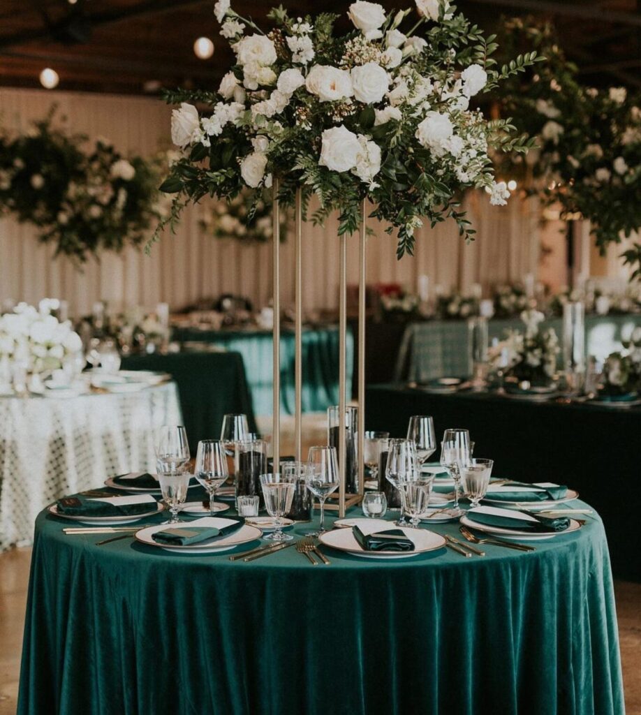 A wedding table set with dark green velvet cloth, white plates, and a tall white floral centerpiece.