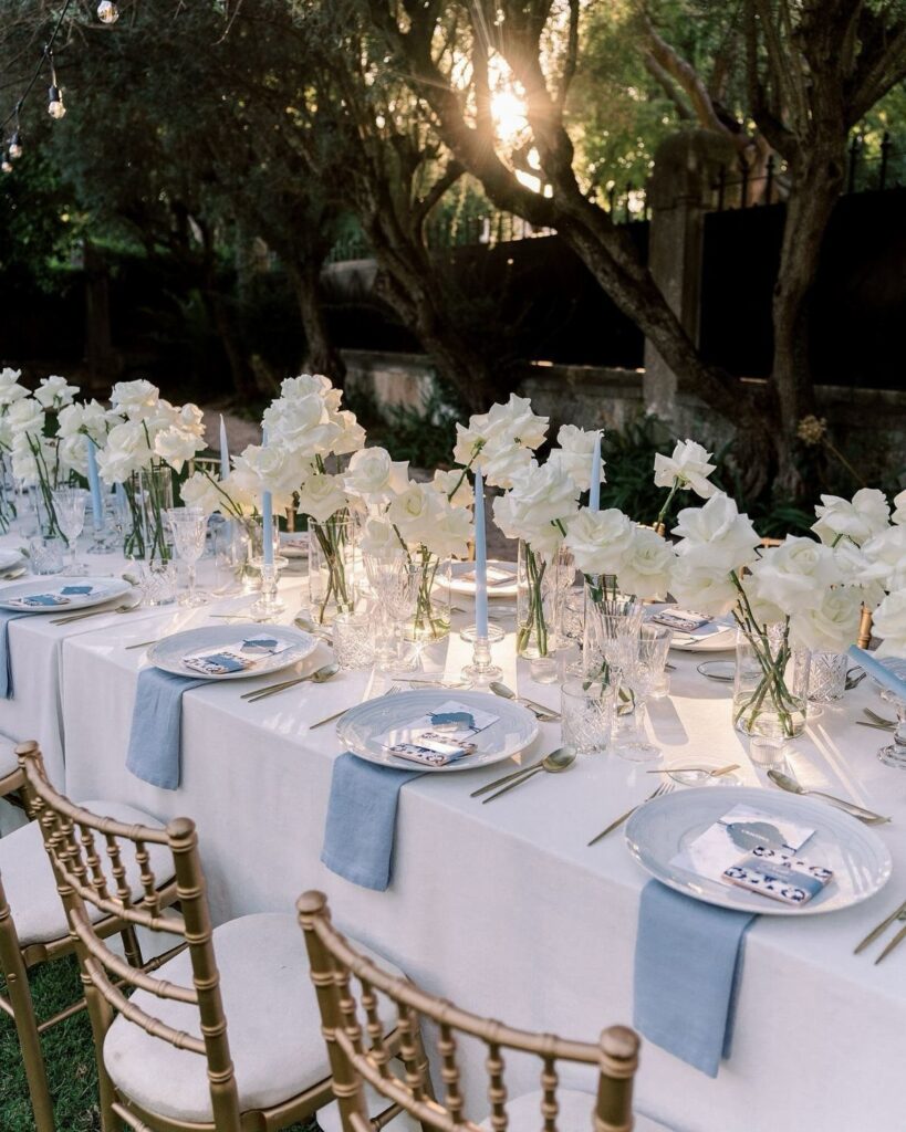 White floral centerpieces and blue napkins on an elegant outdoor dining table setting.