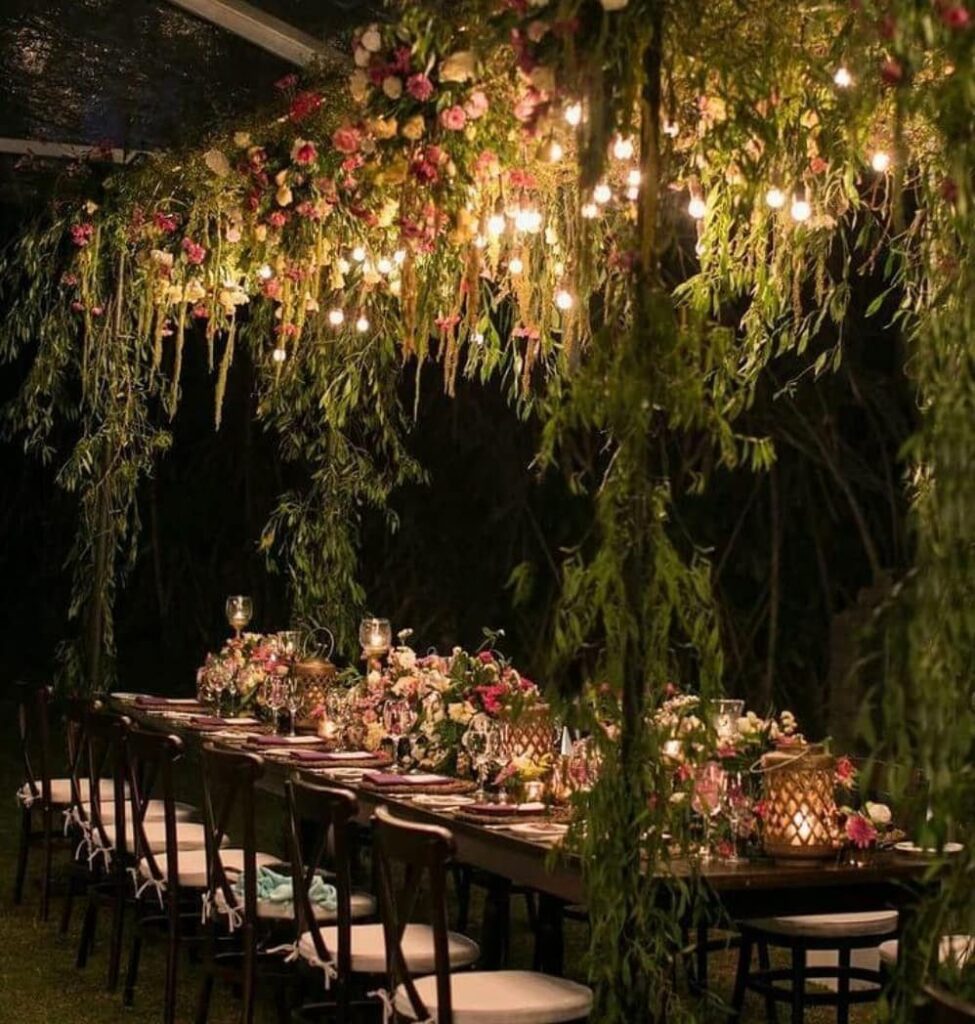 A long dining table decorated with flowers and lights under a canopy of hanging greenery.