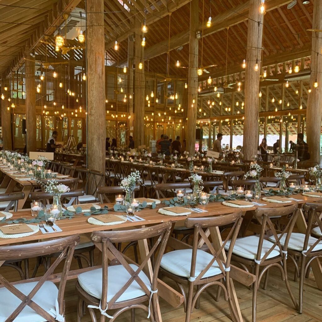 Long wooden banquet tables set with greenery and candles inside a rustic wooden pavilion.