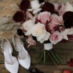 A wedding bouquet with pink and burgundy roses next to white bridal shoes.