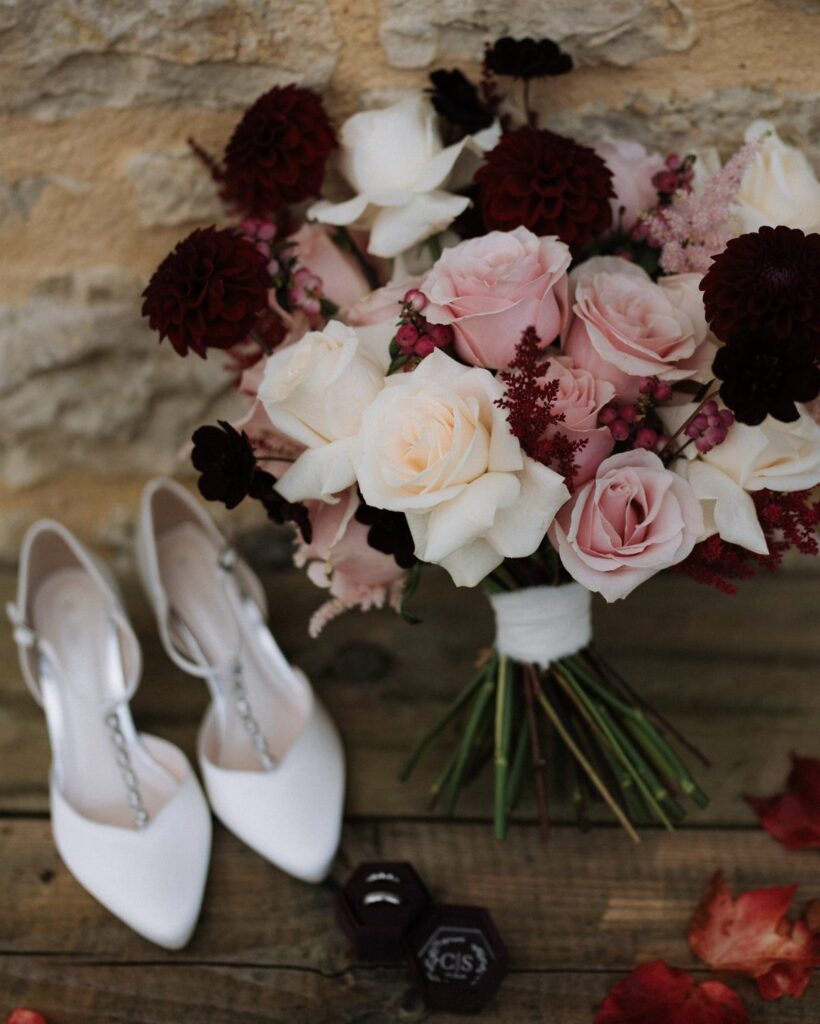 A wedding bouquet with pink and burgundy roses next to white bridal shoes.