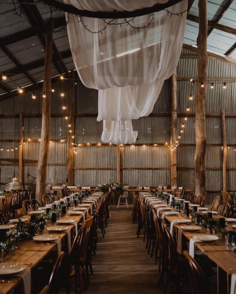 Long wooden banquet tables set for a wedding inside a rustic barn with twinkling lights.