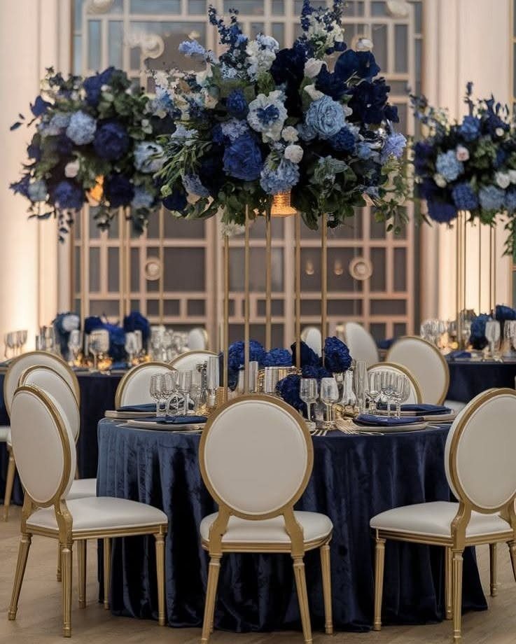 Navy blue velvet tablecloths and large blue floral centerpieces on gold stands at a wedding reception.