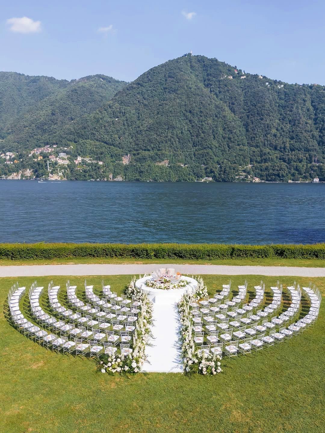 White wedding chairs arranged in semi-circles on a lawn facing a lake and mountains.