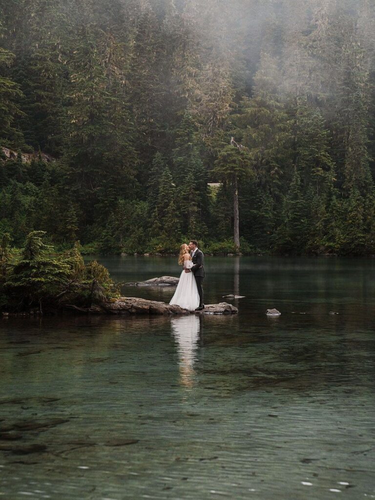 A bride and groom standing on a rock in a misty forest lake.
