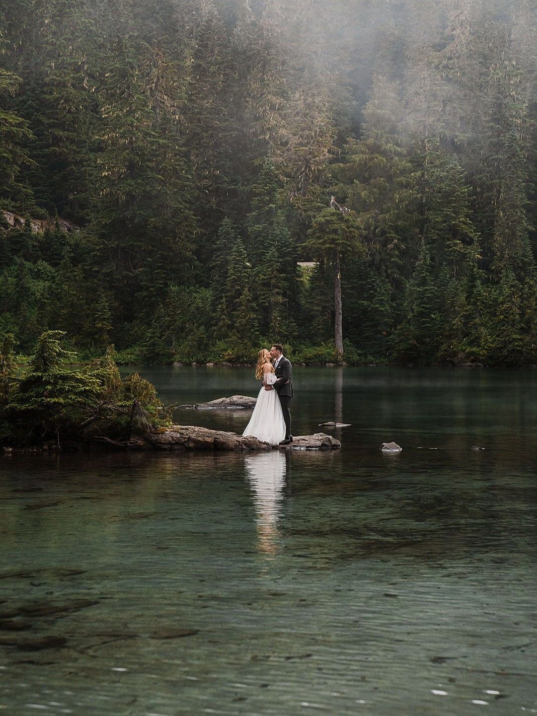 A bride and groom standing on a rock in a misty forest lake.