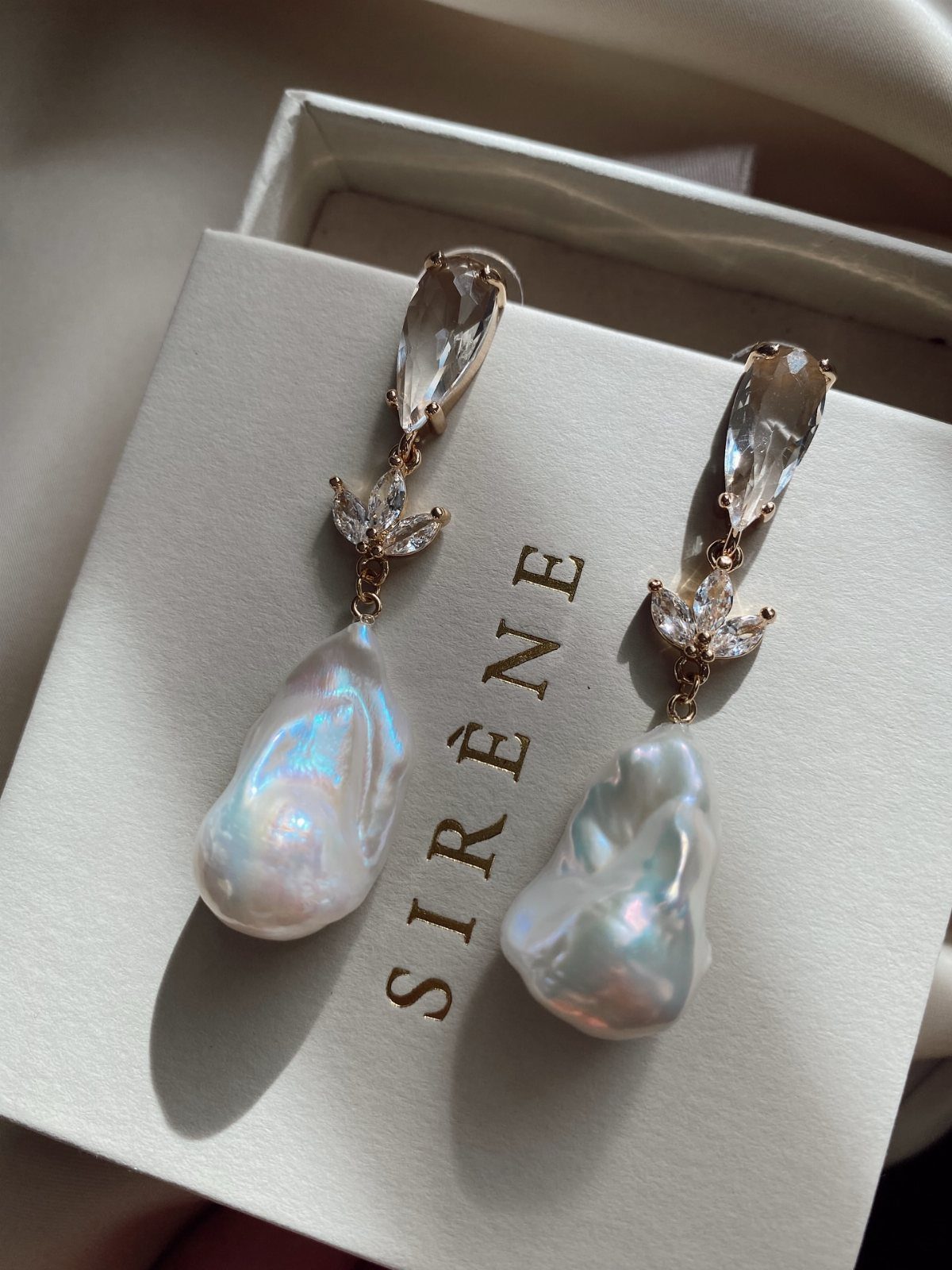 Baroque pearl drop earrings with crystal accents resting on a white brand card.