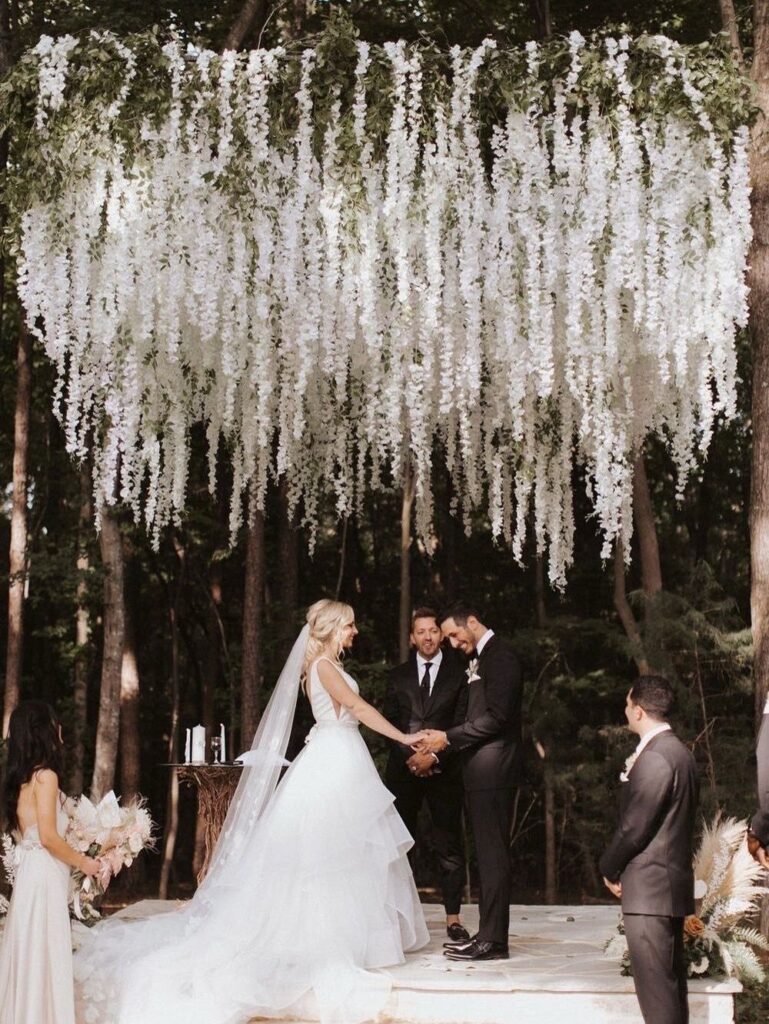A bride and groom hold hands under a large hanging white floral installation in a forest.