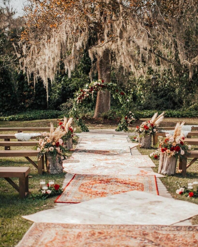 Patterned rugs laid on grass forming a wedding aisle leading to a floral archway.