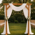 Wooden wedding arch decorated with white fabric drapes and floral bouquets on a green lawn.