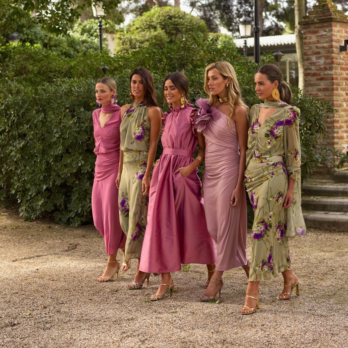 Five women in elegant formal dresses posing together in a lush green garden setting.