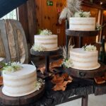 Three white naked cakes with small white flowers on rustic wooden stands atop a wooden table.