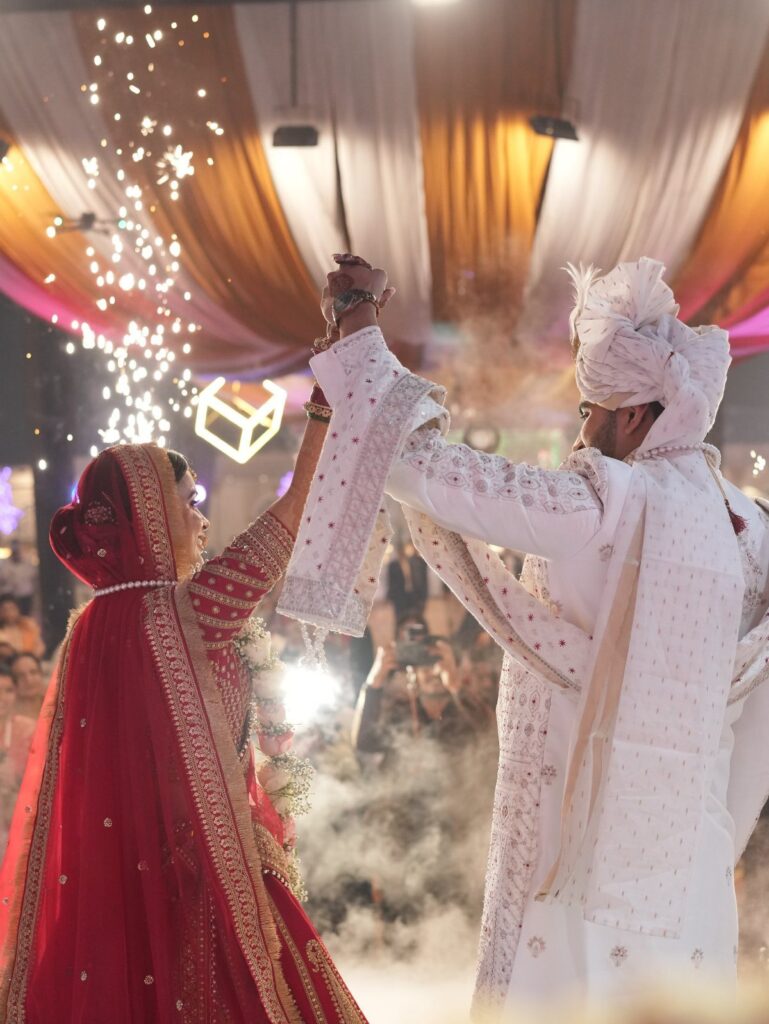 A bride in red and a groom in white dance together amidst smoke and sparkles.