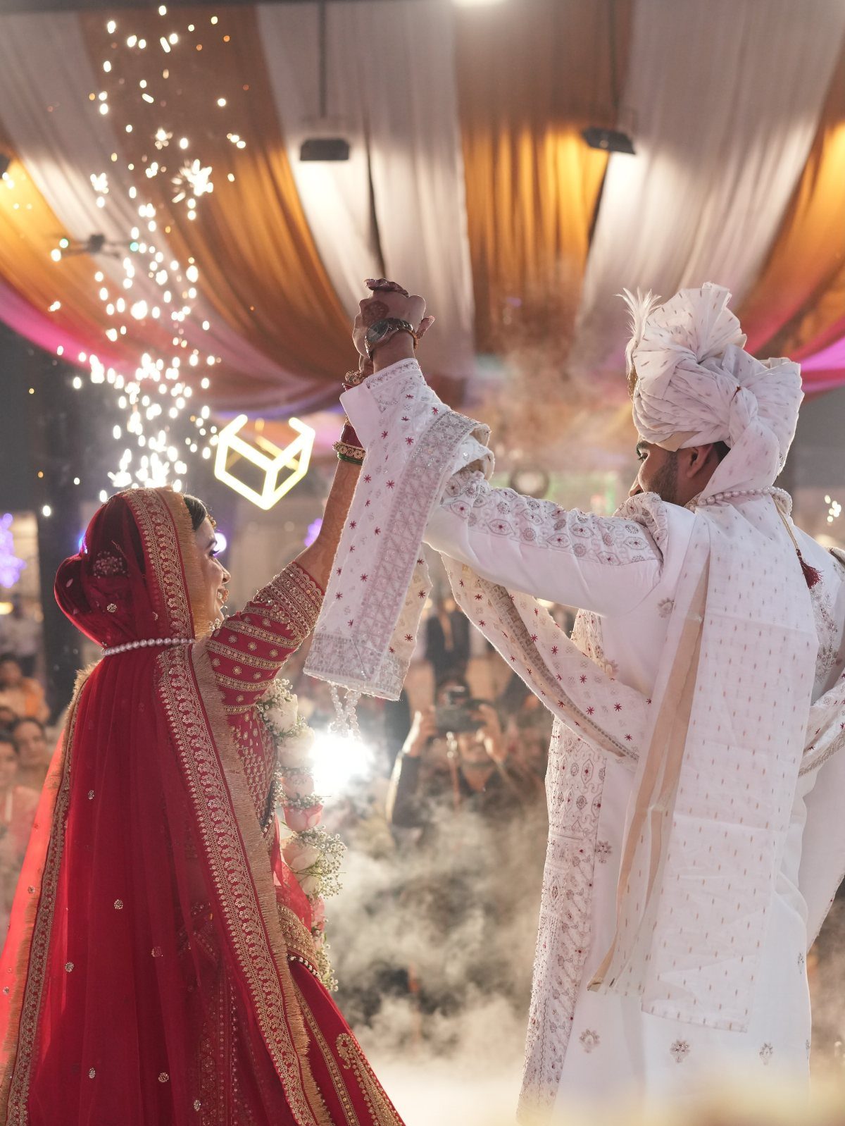 A bride in red and a groom in white dance together amidst smoke and sparkles.