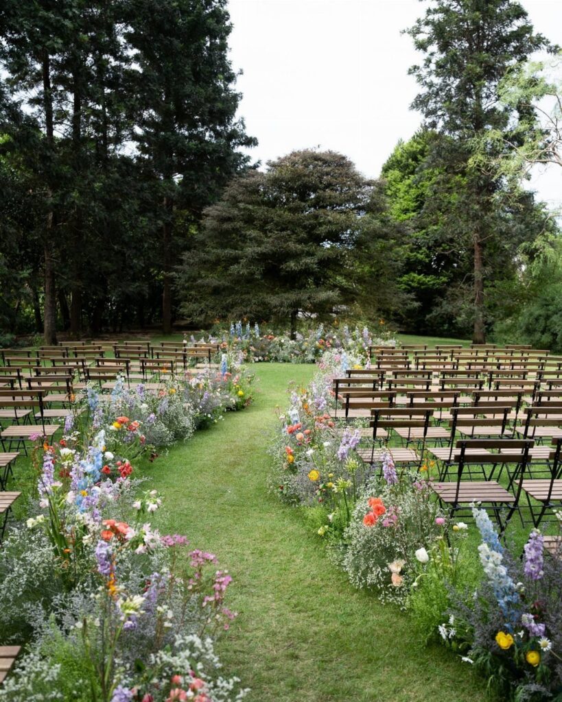Wildflowers line a grassy wedding aisle flanked by rows of wooden chairs under tall green trees.