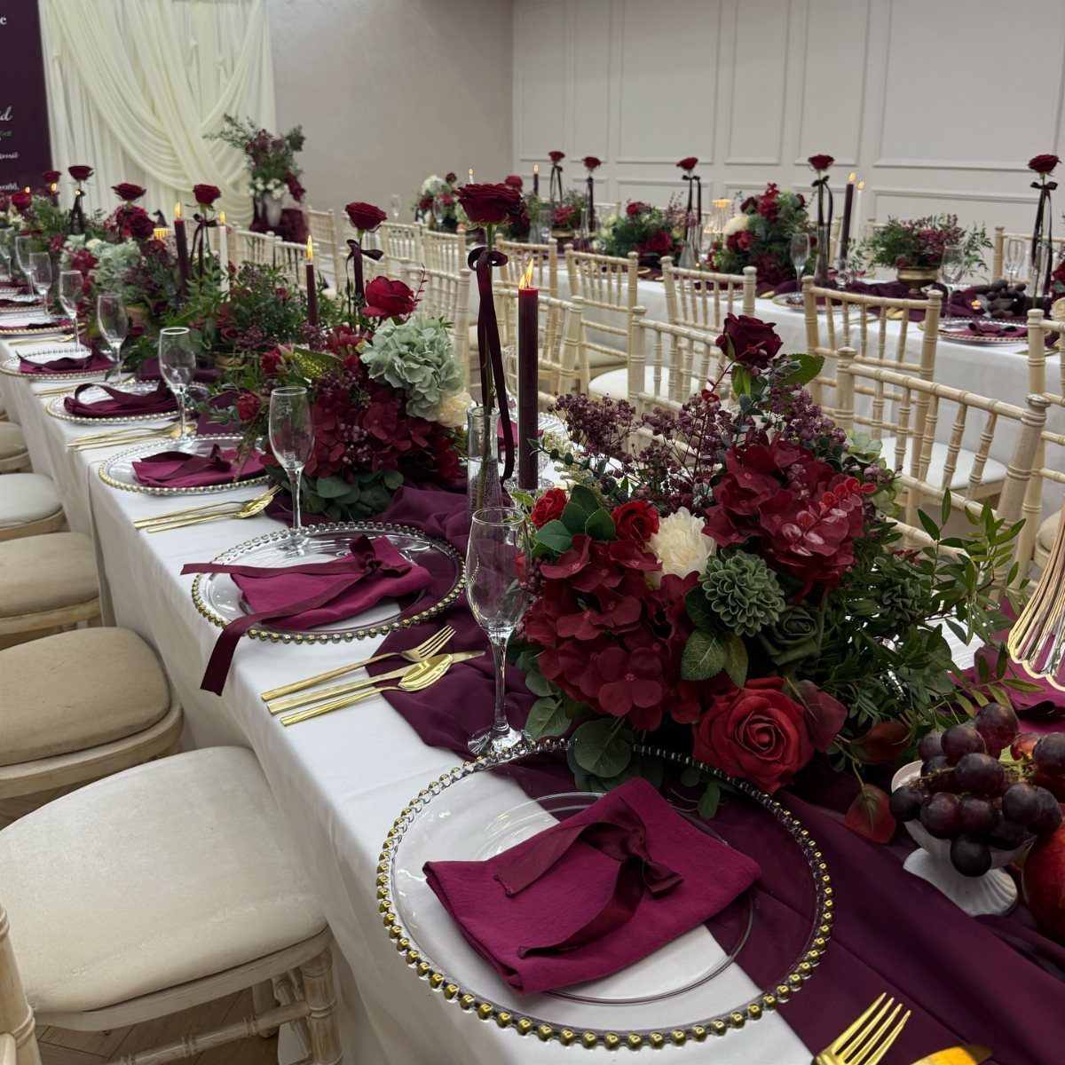 Wedding reception table decorated with burgundy napkins, gold cutlery, and deep red floral centerpieces.